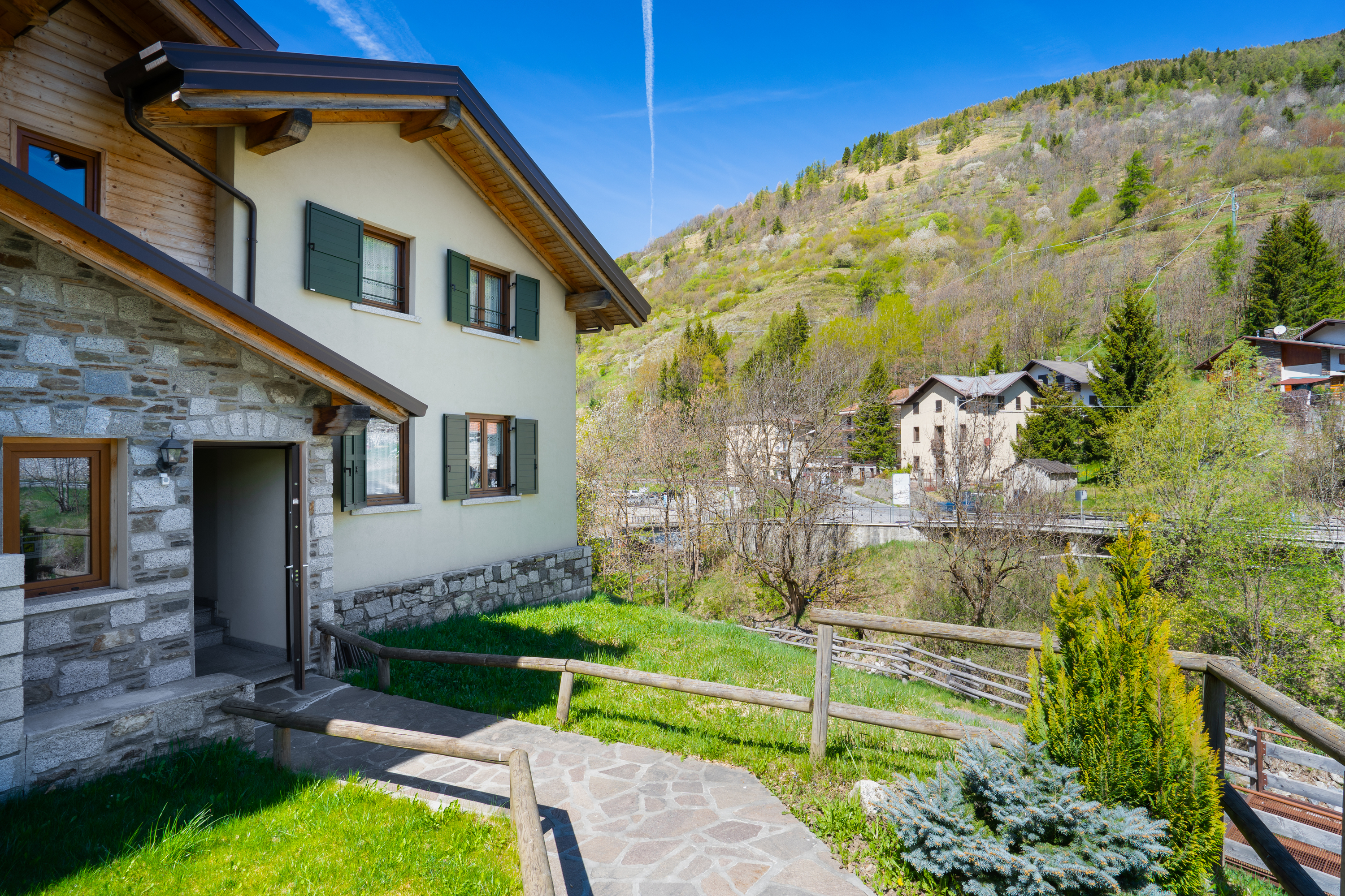Exterior view of a two-story house with stone and stucco siding, green shutters, and a stone walkway leading to the entrance. A grassy yard and mountain backdrop complete the scene.