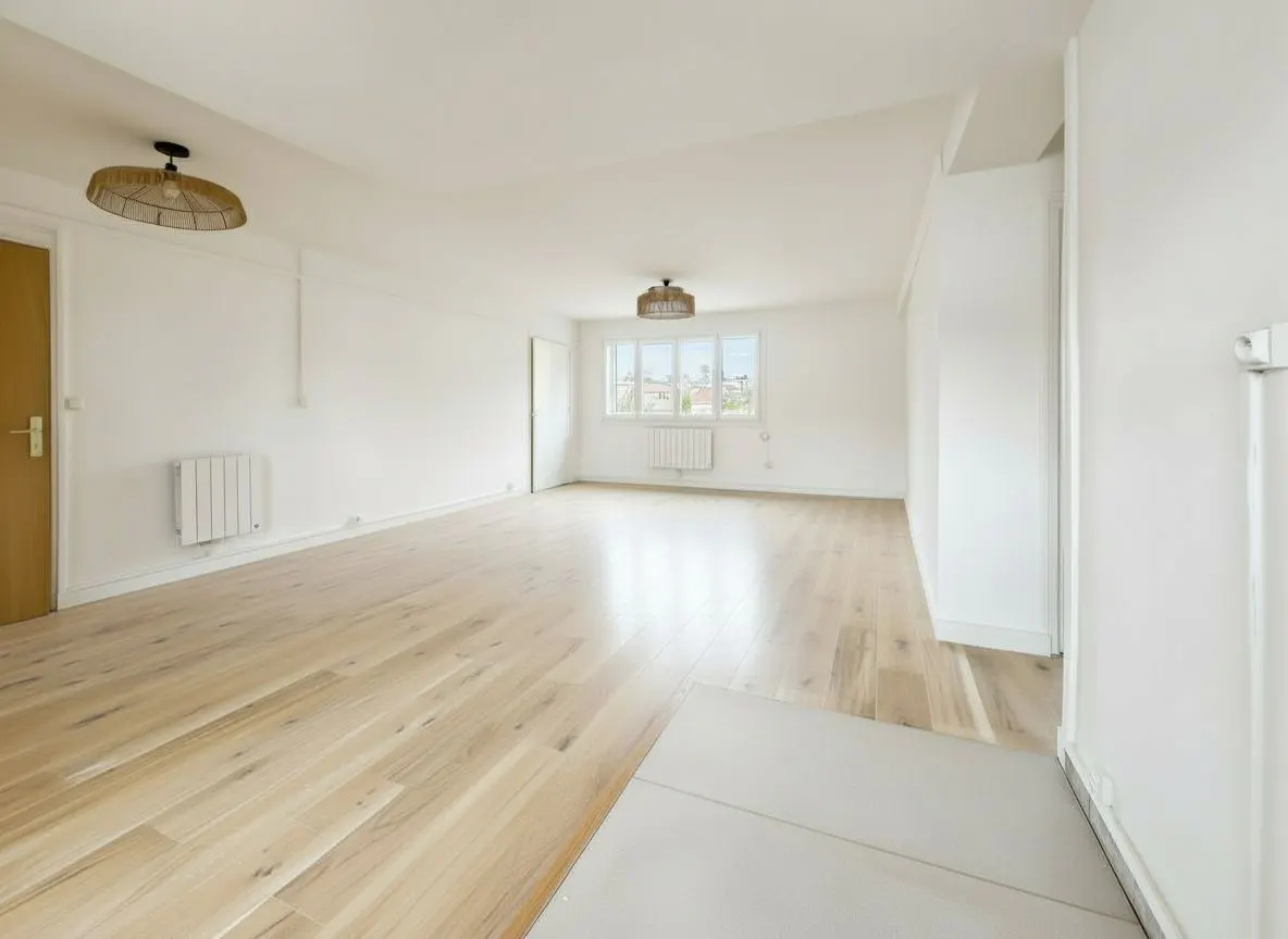 Bright, empty room with light wood floors, white walls, and two straw pendant lights. A window shows a cityscape in the distance.
