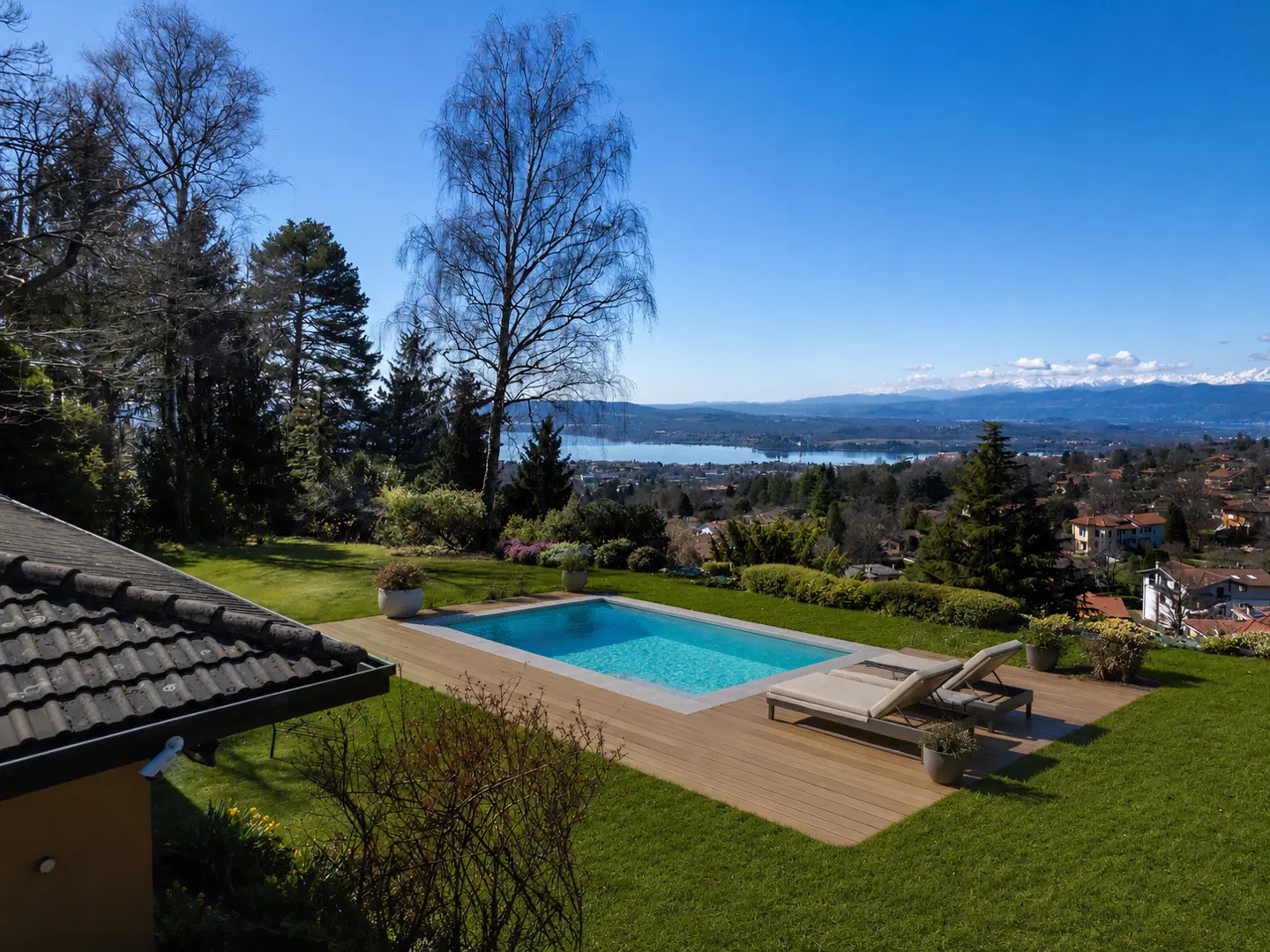 A backyard with a pool, wooden deck, and two lounge chairs overlooking a lake and mountains on a sunny day.