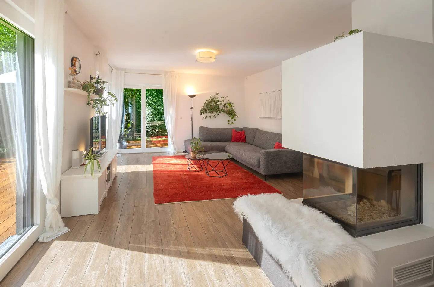 Bright living room with a grey sectional, vibrant red rug, modern fireplace with white fur throw, and glass patio doors.