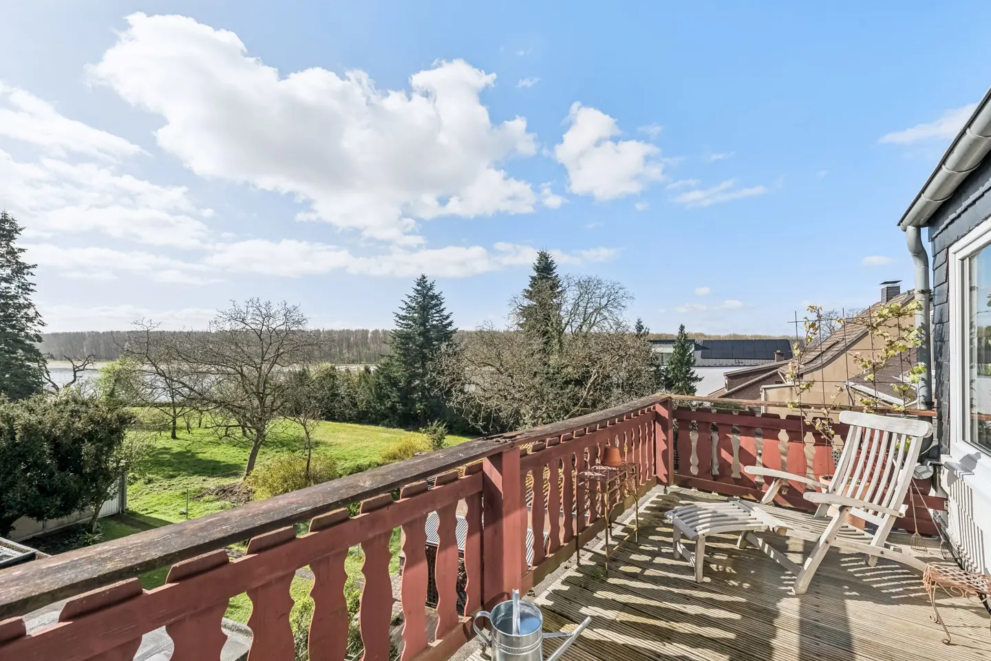 Balcony view with red railing, wooden deck, and white chair. A watering can sits on the deck, overlooking a green lawn and trees under a blue sky.