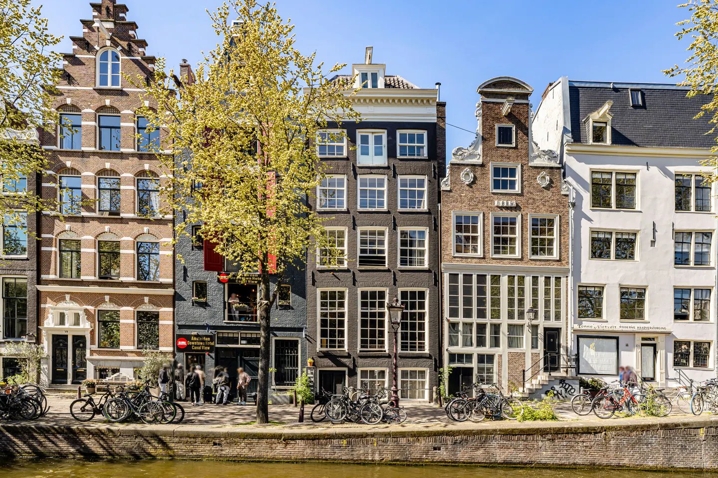 Amsterdam canal view with colorful, narrow houses, bikes parked along the brick canal wall, and a tree with yellow leaves.