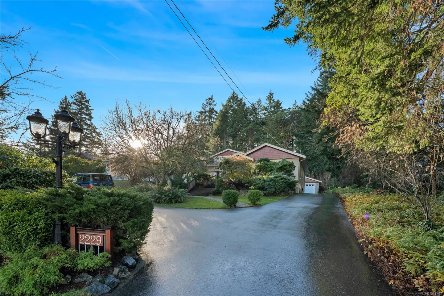 Exterior view of a tan house with a red roof, surrounded by trees and greenery, under a blue sky. A driveway leads to the house. The address "2229" is visible on a sign.