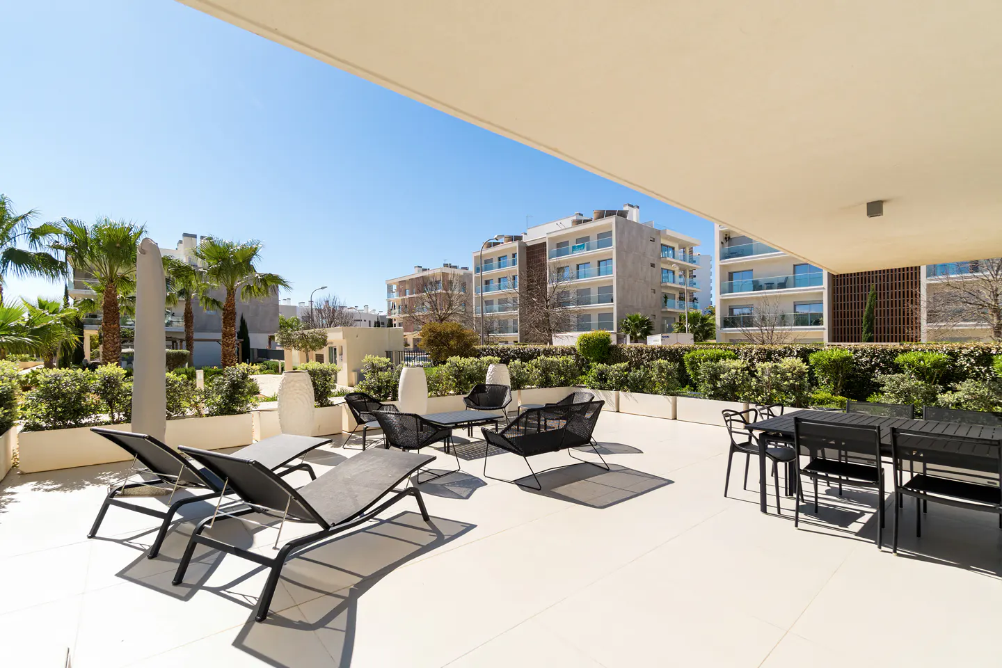 Outdoor patio with black lounge chairs and dining set on a white tiled floor, with palm trees and buildings in the background.