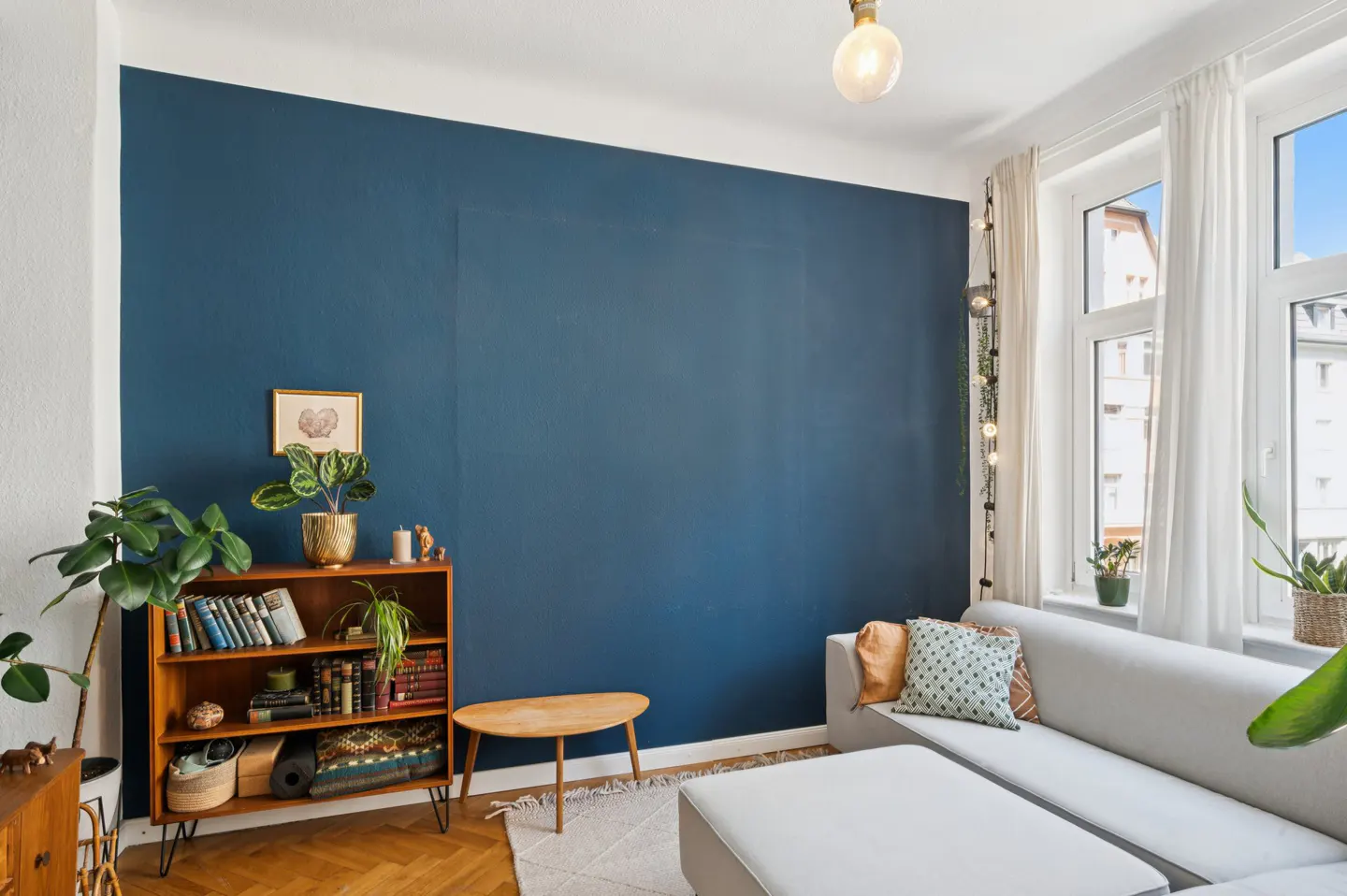 Living room with a blue accent wall, a white sofa, a wooden bookshelf, and a herringbone floor. Natural light streams through the window.