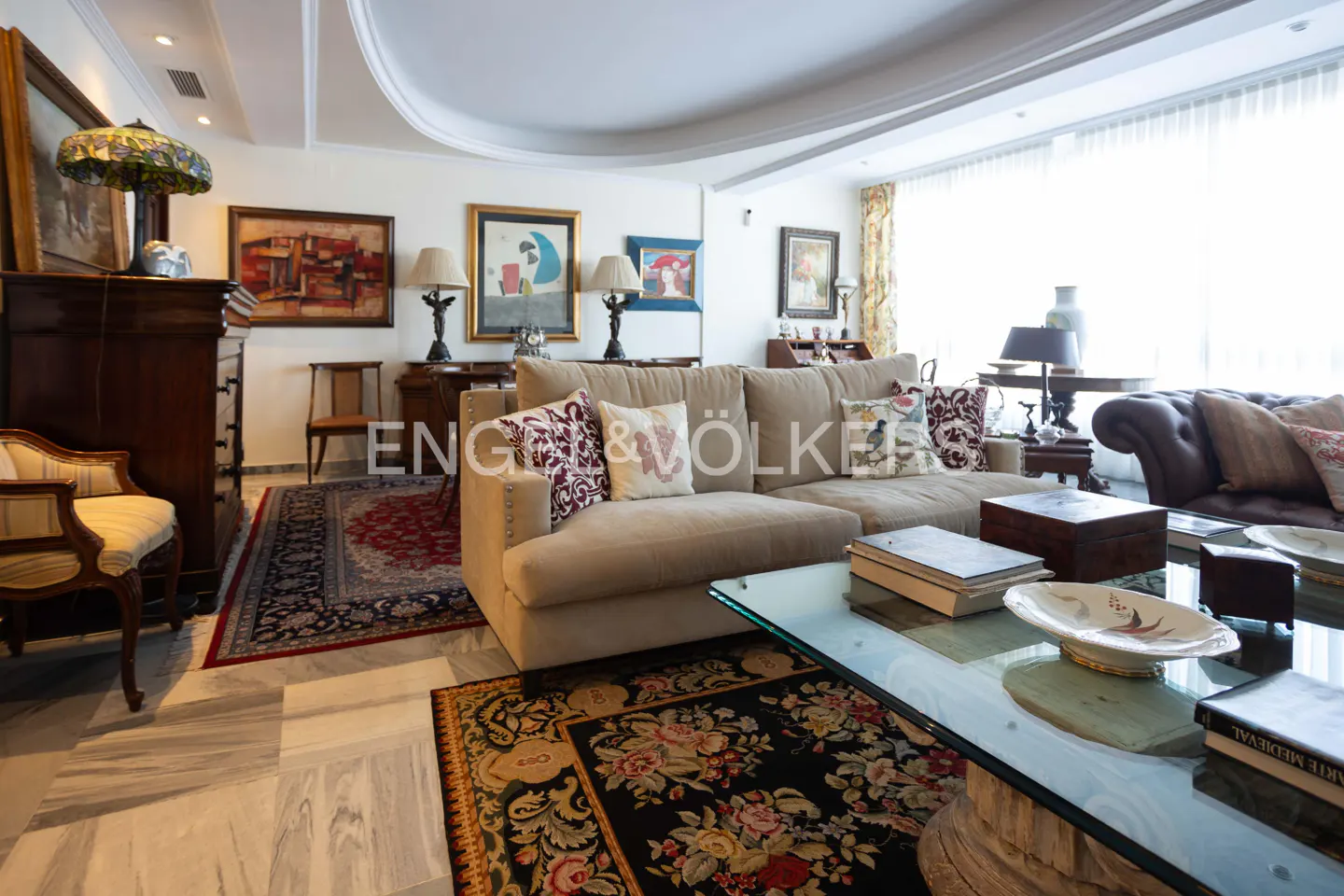 A bright living room with beige sofas, floral rugs, and marble floors. Art hangs on the walls, and a glass coffee table sits in the center.