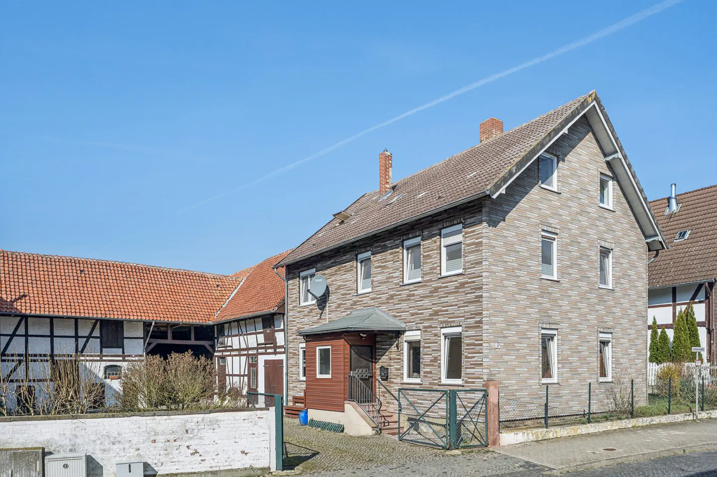 Two-story brick house with a brown roof and white-framed windows under a clear blue sky. A half-timbered building is visible in the background.