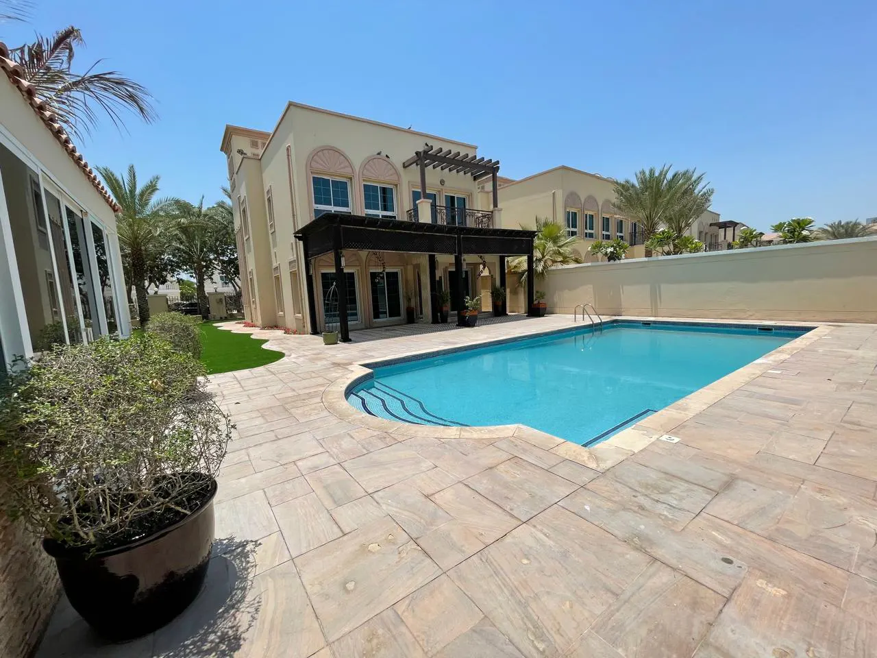 A beige two-story house with a blue swimming pool and a stone patio under a clear blue sky.