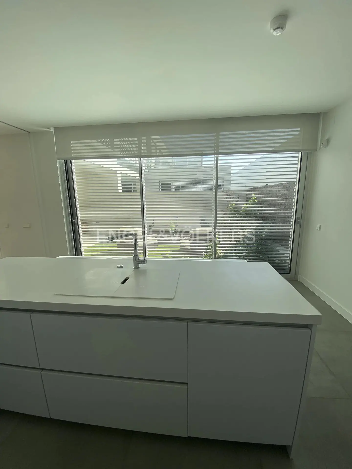 A modern white kitchen island with a sink and faucet, set against a large window with blinds overlooking a green yard.