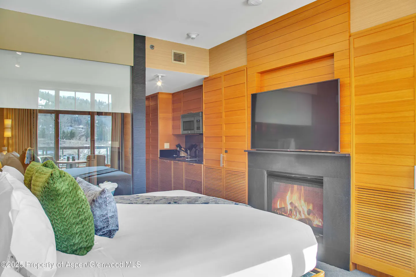 Hotel room with a king bed, green pillows, fireplace, TV, and kitchenette. Large windows overlook a snowy landscape.