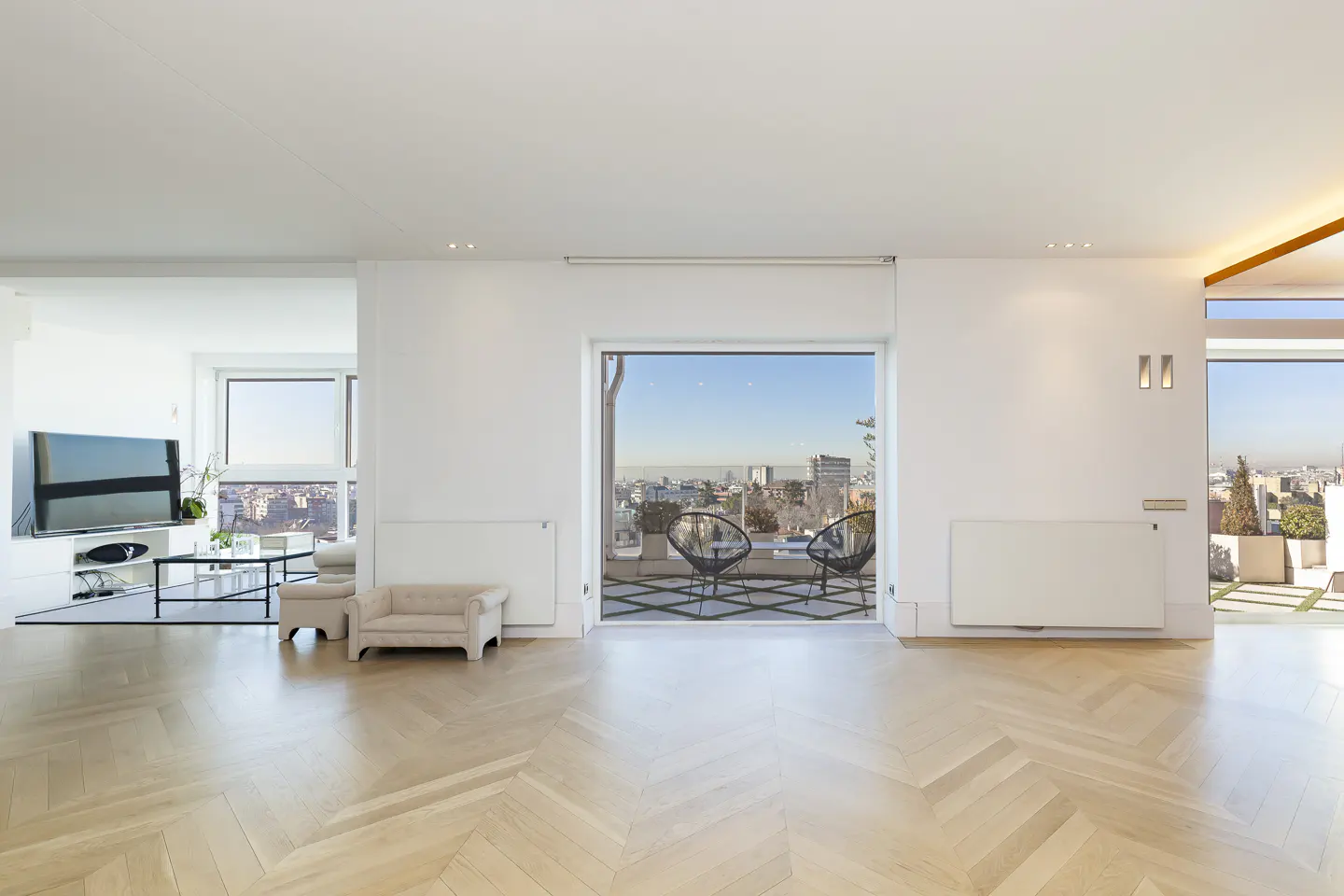 Bright, modern apartment interior with herringbone wood floors, white walls, and a balcony with city views.