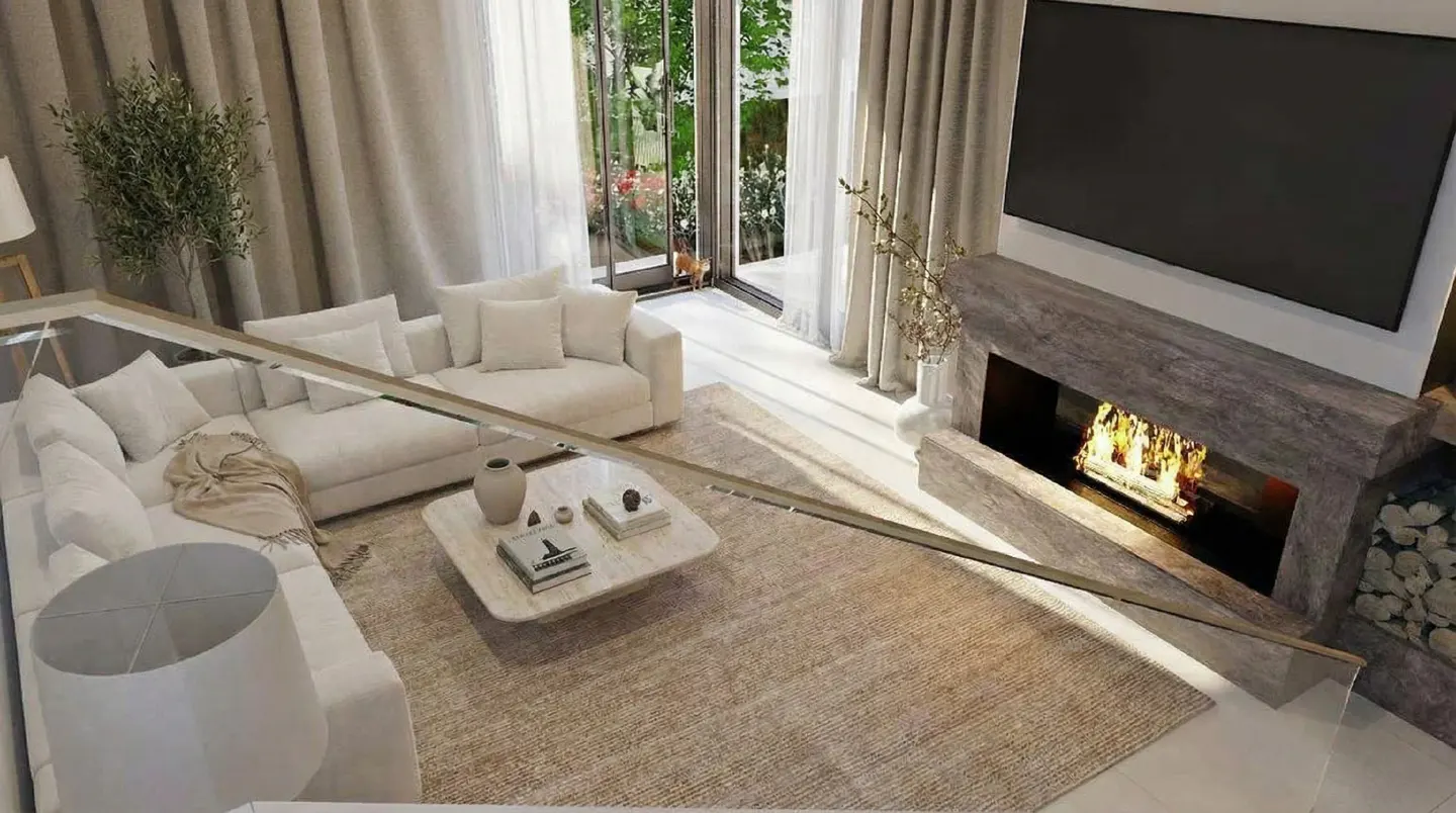 Living room with white sofas, a stone fireplace with a TV above, and a beige rug. Large windows offer a view of greenery.