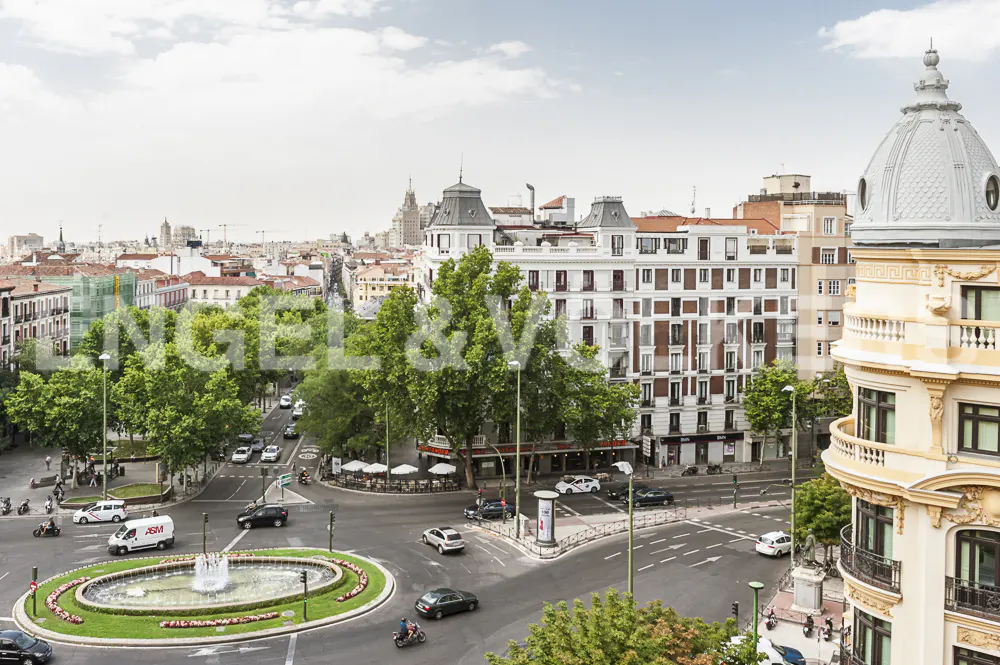 Cityscape view of Madrid, Spain, featuring a roundabout with a fountain, classic architecture, and busy streets under a cloudy sky.