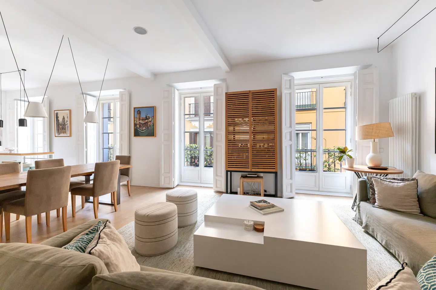 Bright, modern living room with white walls, wood floors, and large windows. A dining table, sofas, and a white coffee table furnish the space.