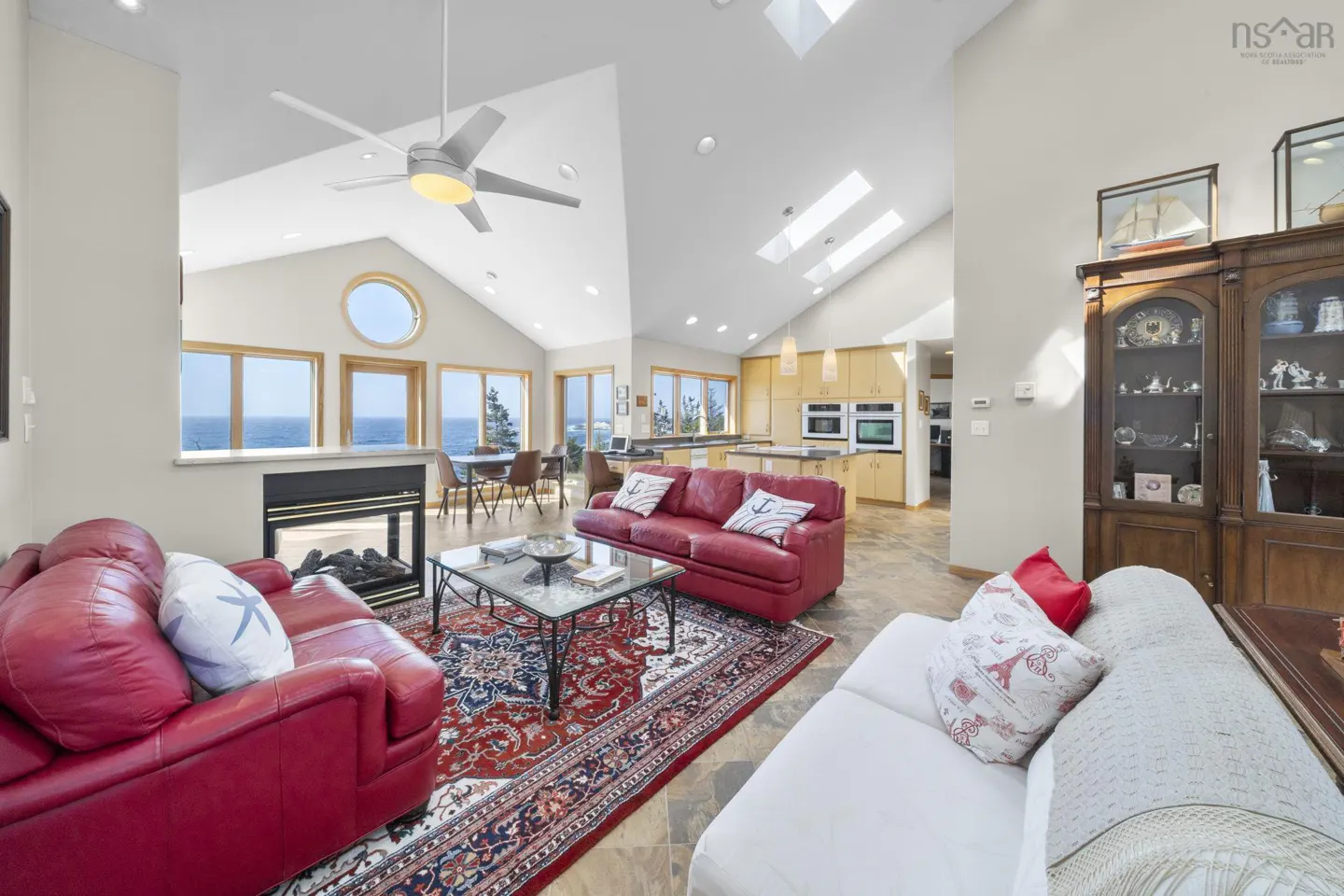 Bright living room with red sofas, patterned rug, and ocean views through large windows. A modern kitchen is visible in the background.