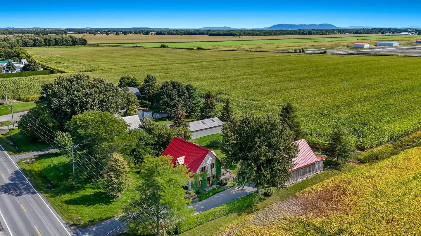 Aerial view of a green house with a red roof, surrounded by trees and fields under a blue sky.