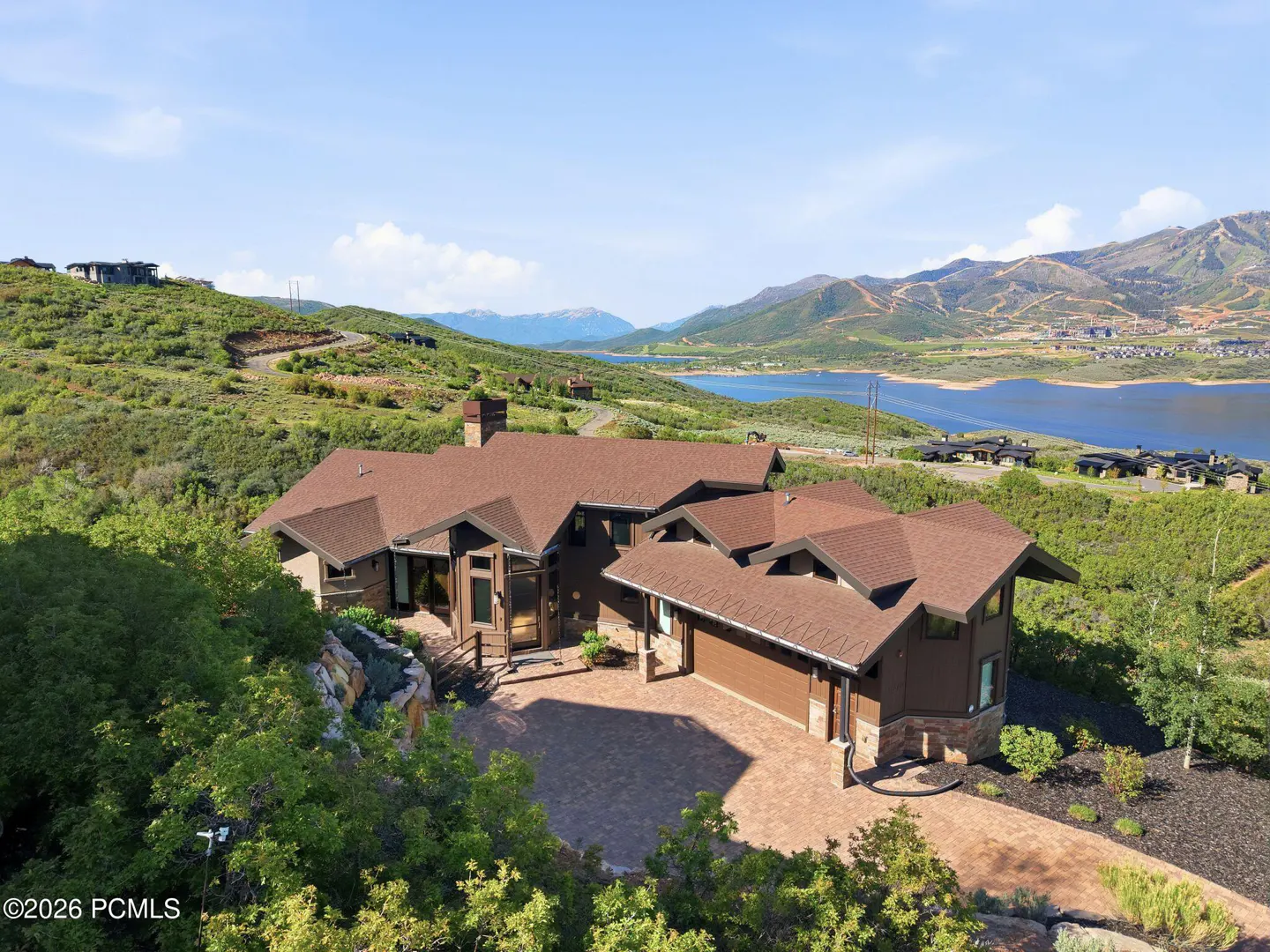 Aerial view of a brown mountain home with a paved driveway, surrounded by lush greenery, a lake, and distant mountains.