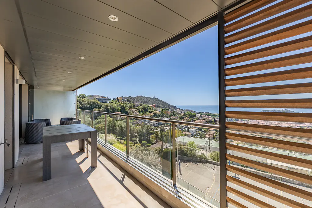 Balcony view with table, chairs, and glass railing overlooking a cityscape with trees and the ocean under a clear blue sky.