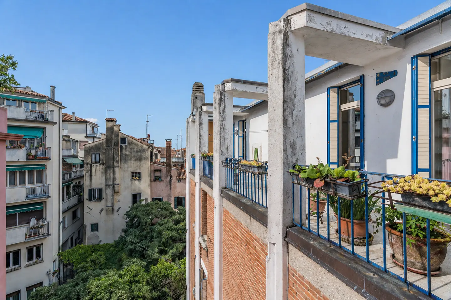 View from a balcony with blue railings and potted plants, overlooking buildings in Venice, Italy, under a clear blue sky.