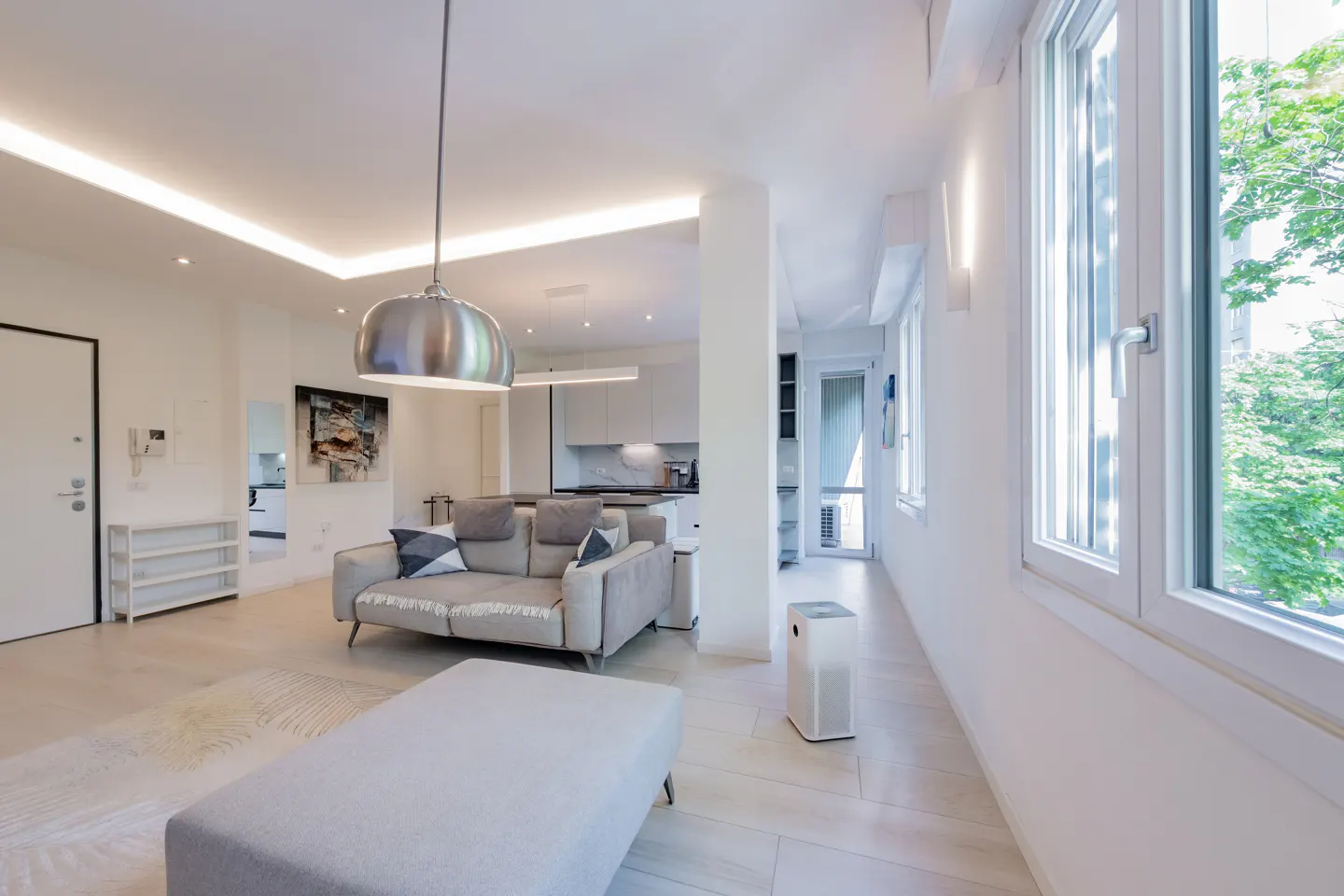 Bright, modern apartment interior with a gray sofa, ottoman, and pendant light. Large windows offer a view of green trees.