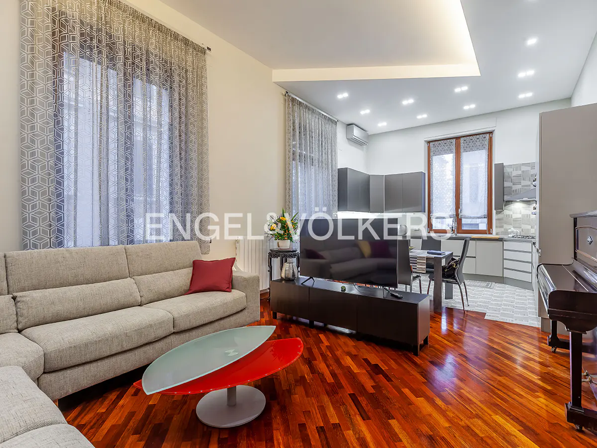 Bright living room with hardwood floors, a gray sofa, a red and gray coffee table, and a TV. The kitchen is visible in the background.