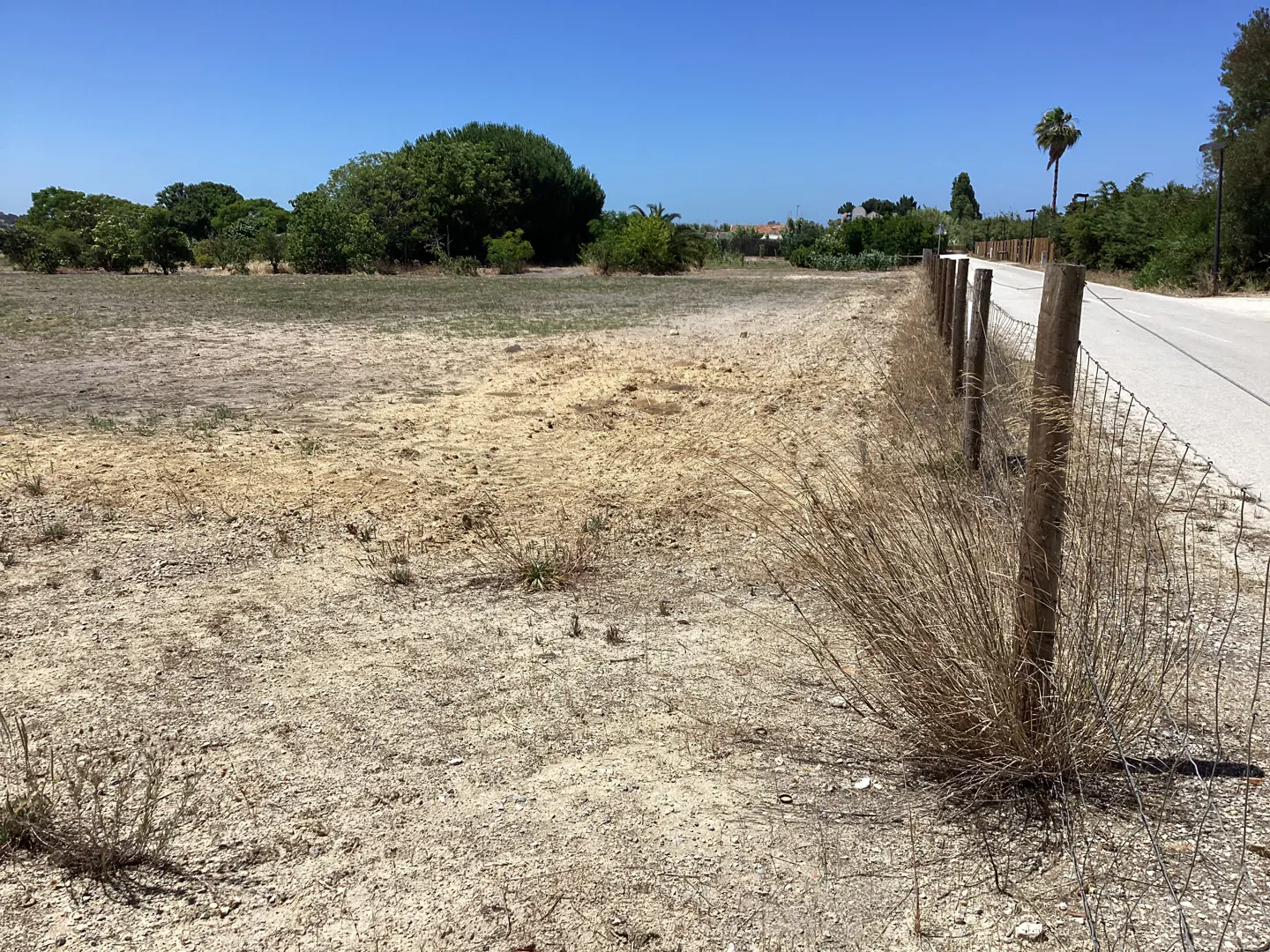 A vacant, dry dirt lot bordered by a wire fence and a paved road under a clear blue sky.