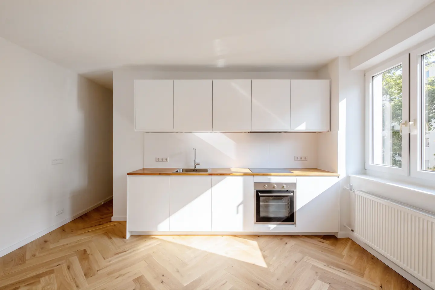 Bright, modern kitchen with white cabinets, wood countertop, and herringbone wood floor. Sunlight streams through a window.