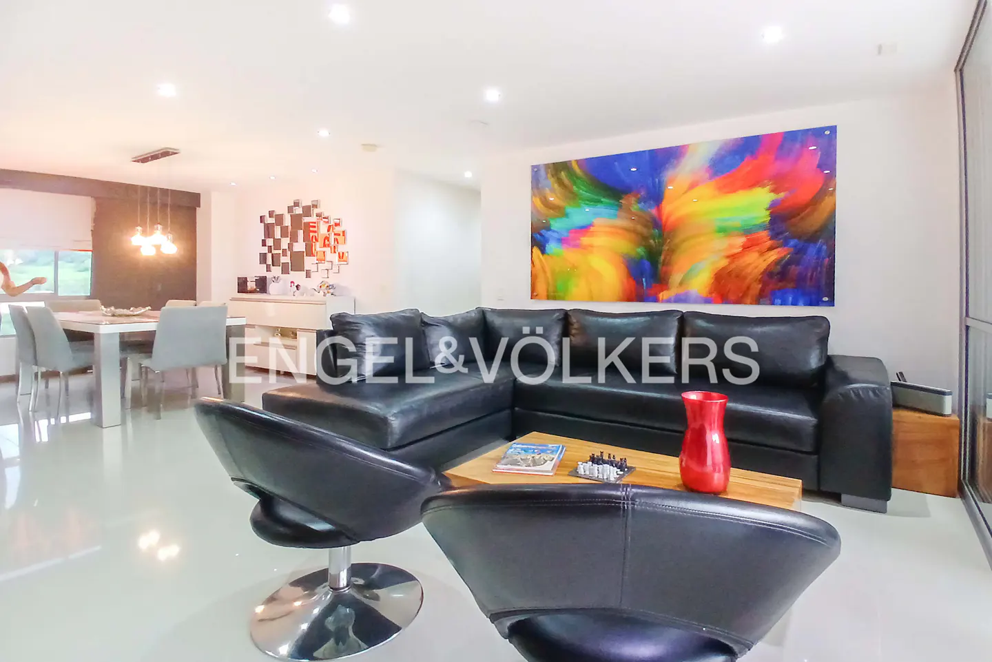 Living room with black leather sofa, chairs, wood table, and colorful abstract art. Dining table in background. Engel & Völkers logo.