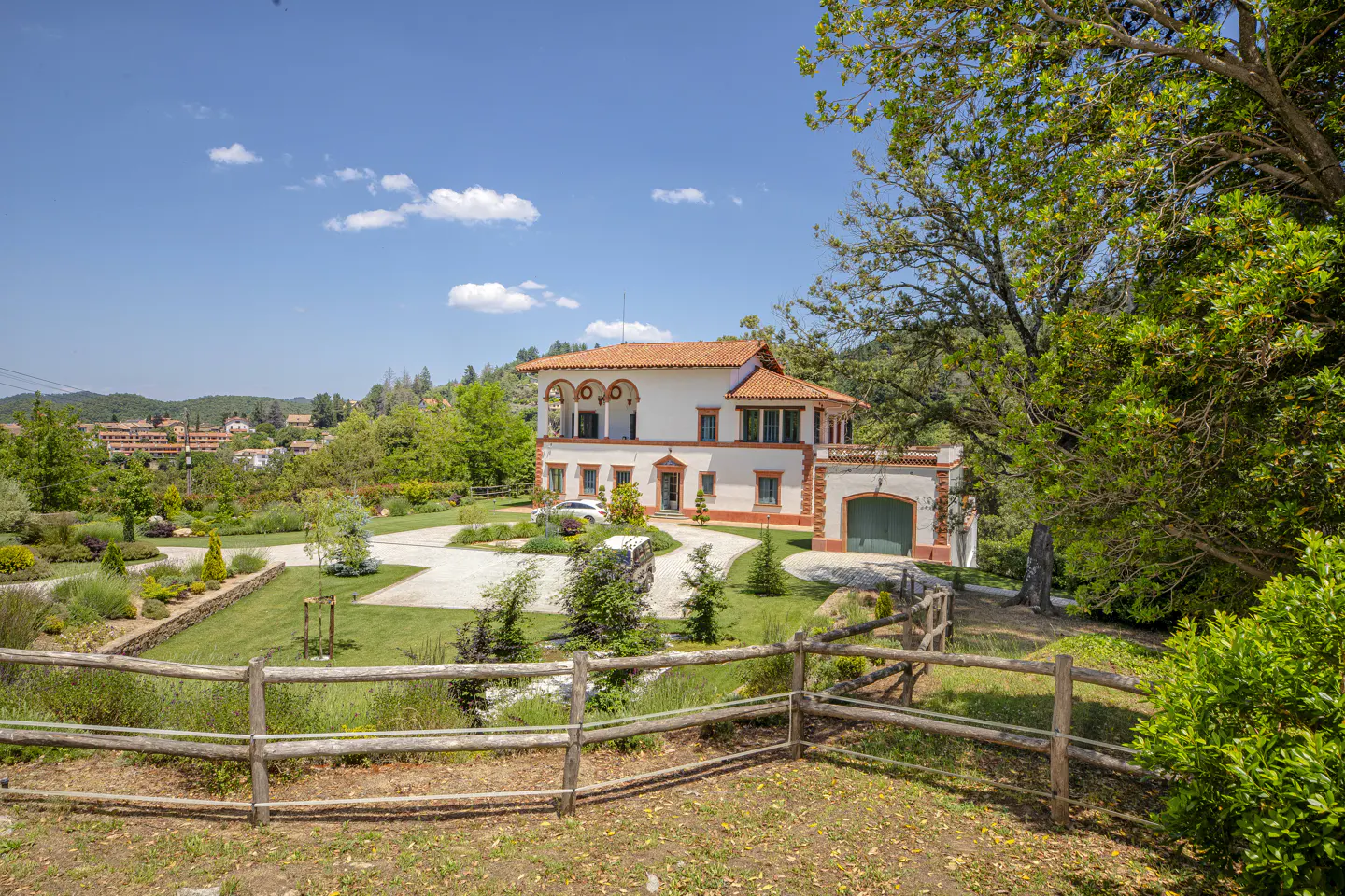 A two-story Mediterranean-style villa with a terracotta roof, white walls, and a green garage door in a sunny landscape.