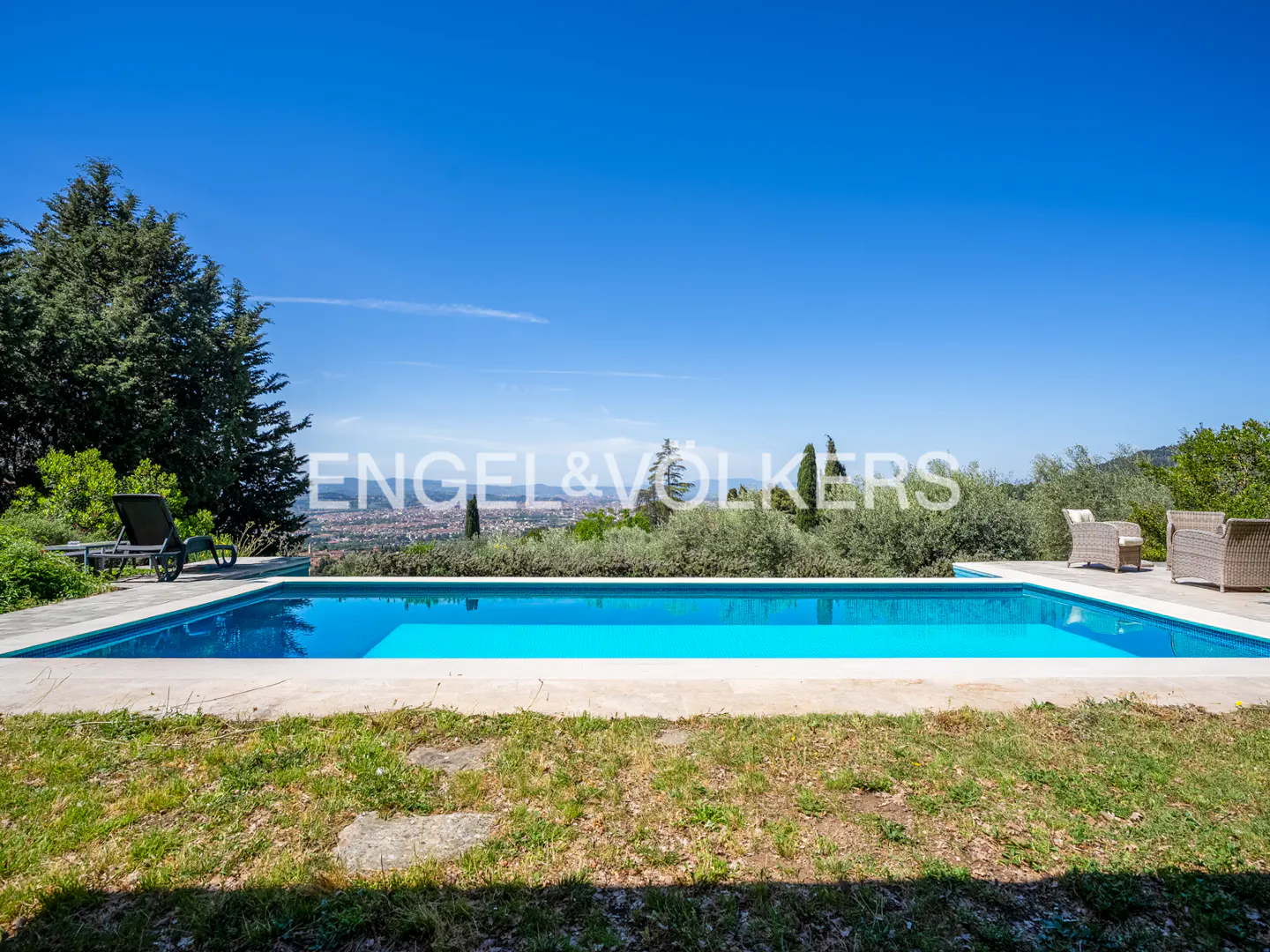 Outdoor pool with blue water, surrounded by green grass and trees, under a clear blue sky. Lounge chairs and wicker furniture are visible.