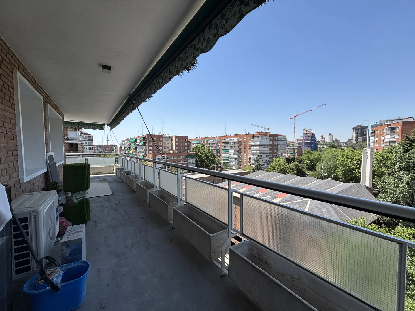 Spacious balcony with a metal railing, planters, and an air conditioning unit, overlooking a sunny urban neighborhood.