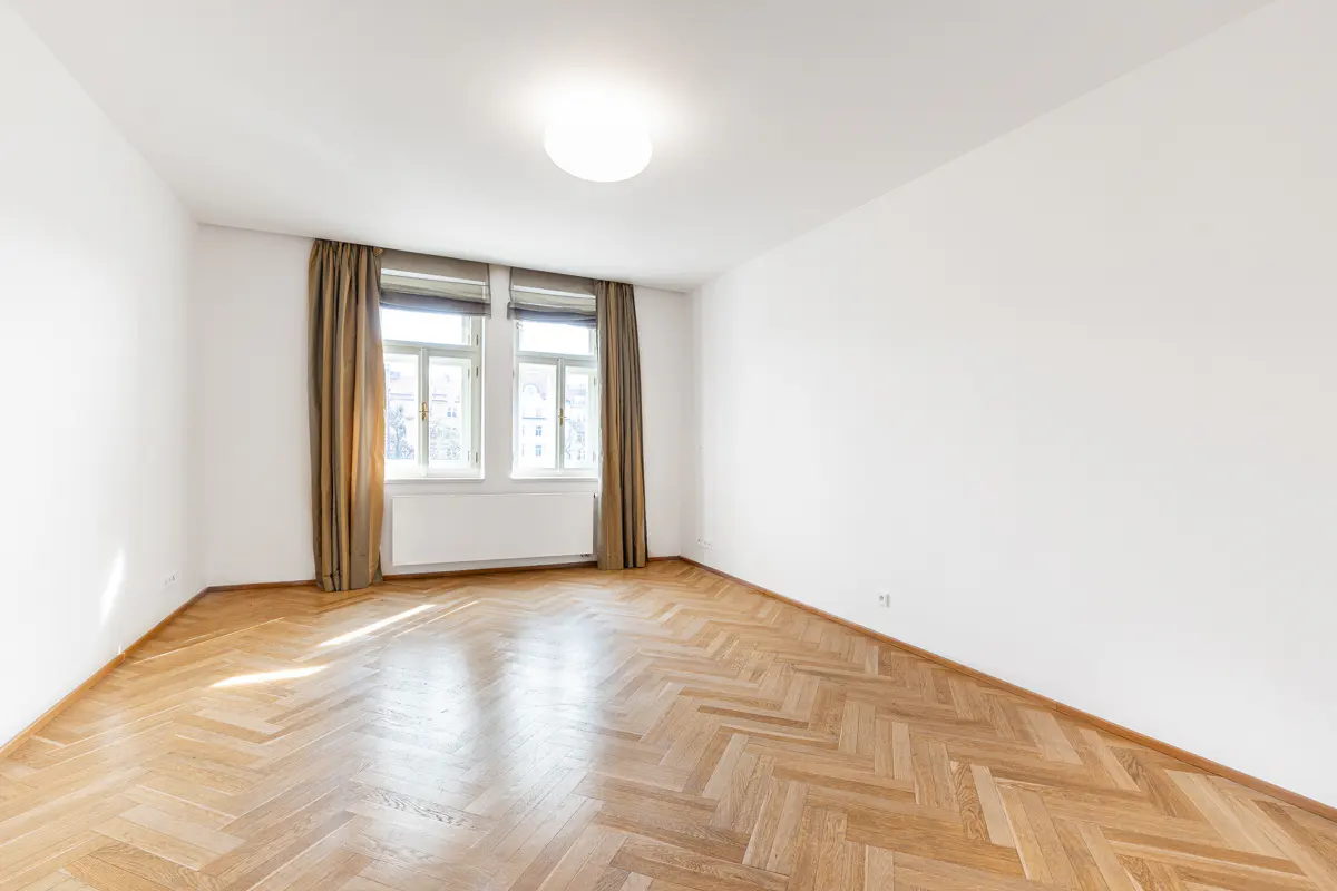 Empty room with white walls, herringbone wood floor, two windows with curtains, and a ceiling light.