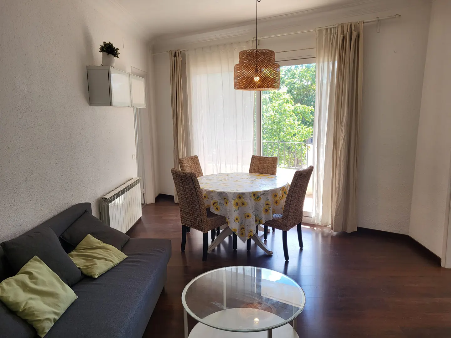 Bright living room with a gray sofa, round table with sunflower tablecloth, and wicker chairs. A woven pendant light hangs above.