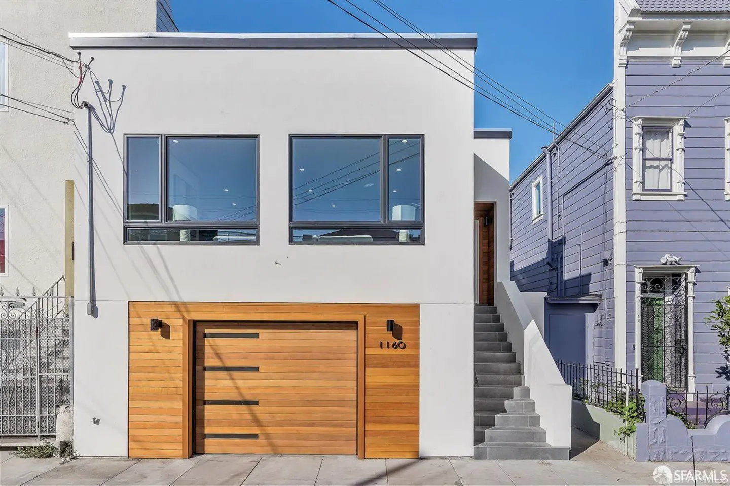 Modern white house with a wooden garage door and gray stairs leading to the front door. A blue house is visible on the right.