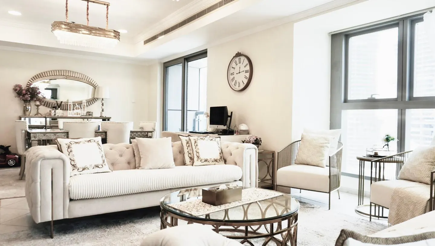 A bright living room with a cream sofa, chairs, and a glass-topped coffee table. A large window and a wall clock are visible.