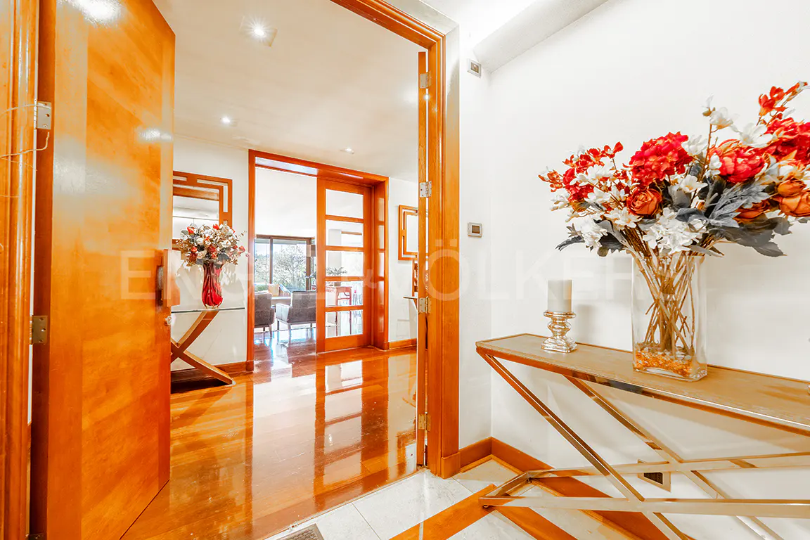 Open wooden door reveals a bright hallway with hardwood floors. A console table with flowers sits against a white wall.
