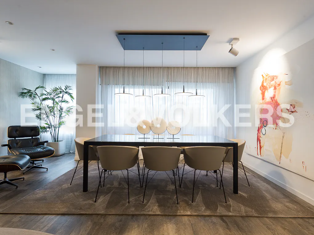 A modern dining room features a black table with six beige chairs, a large abstract painting, and a black leather lounge chair.