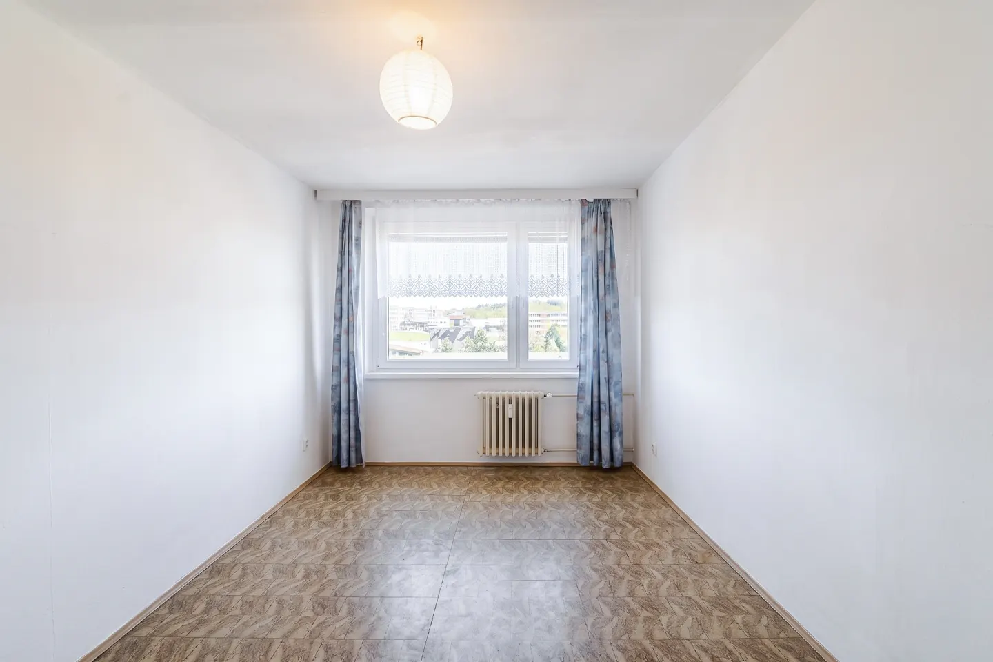 Empty room with white walls, patterned brown flooring, a central window with blue curtains, and a ceiling light fixture.