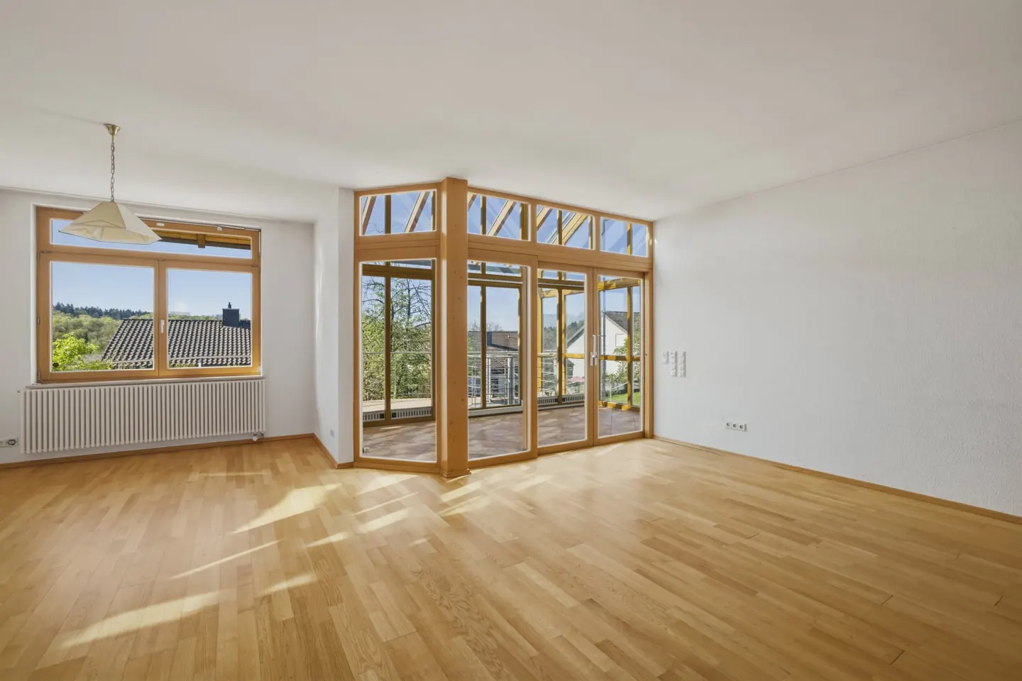 Bright, empty room with wood floors, white walls, and a large window with a view. A glass-enclosed sunroom is adjacent.