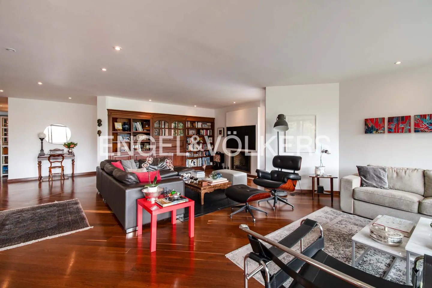 Spacious living room with hardwood floors, leather sofas, a bookcase, and modern furniture. The walls are white, and there are colorful paintings.