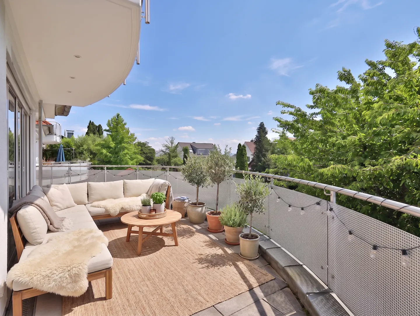 A bright, sunny balcony with a beige sectional sofa, round wooden table, potted plants, and a woven rug. Trees and a blue sky are visible in the background.