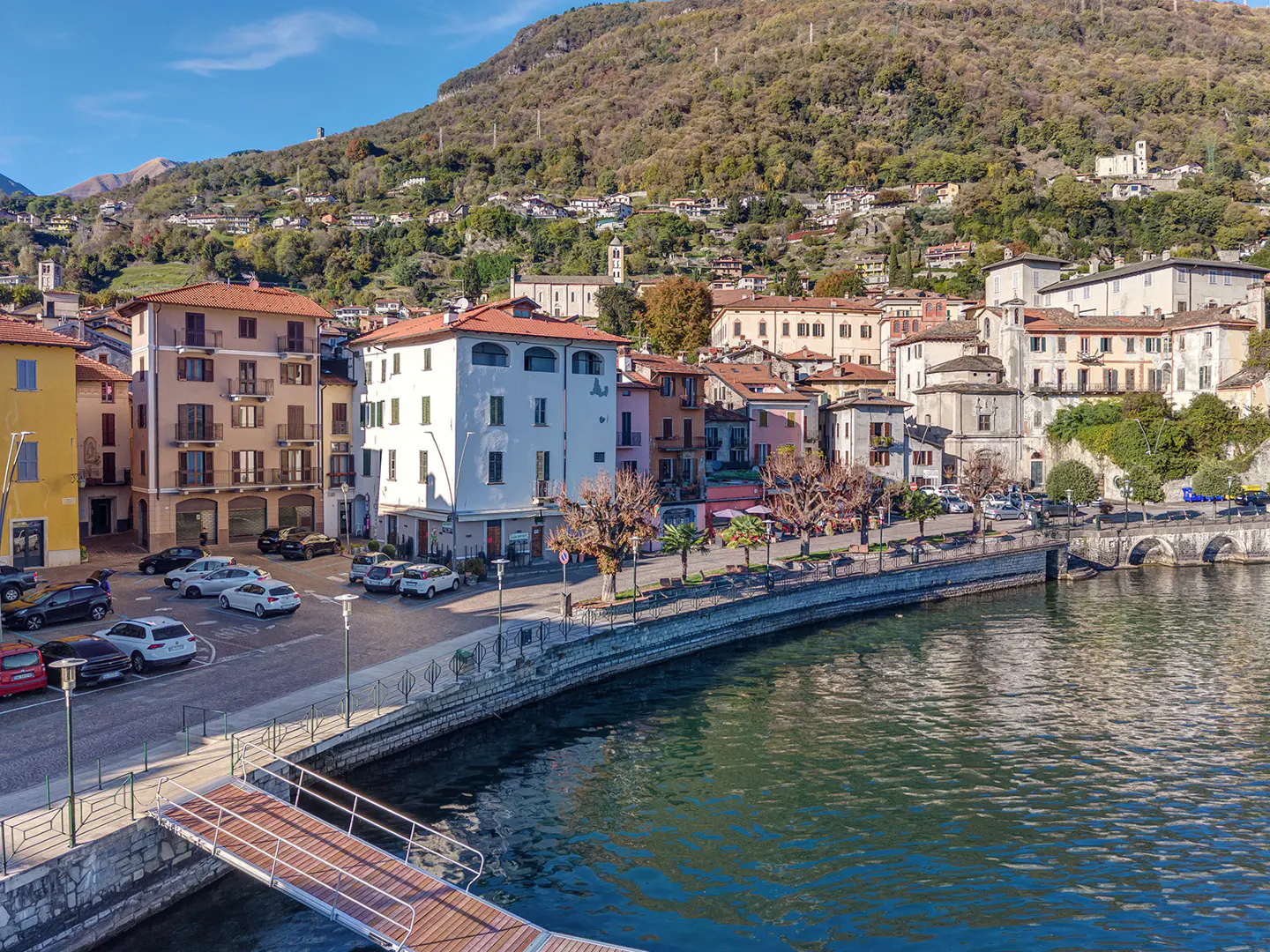 Scenic view of Argegno, Italy, featuring colorful buildings, a waterfront promenade, and a mountain backdrop under a clear blue sky.