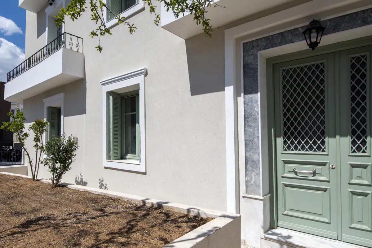Exterior view of a two-story house with a light green door and window shutters. A small balcony is visible on the second floor.