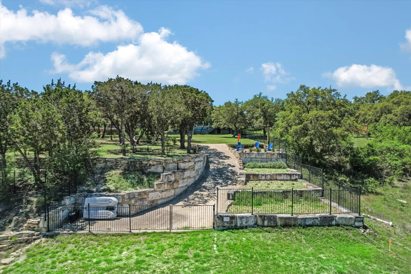 Aerial view of a landscaped yard with stone walls, black metal fences, and green trees under a blue sky with white clouds.