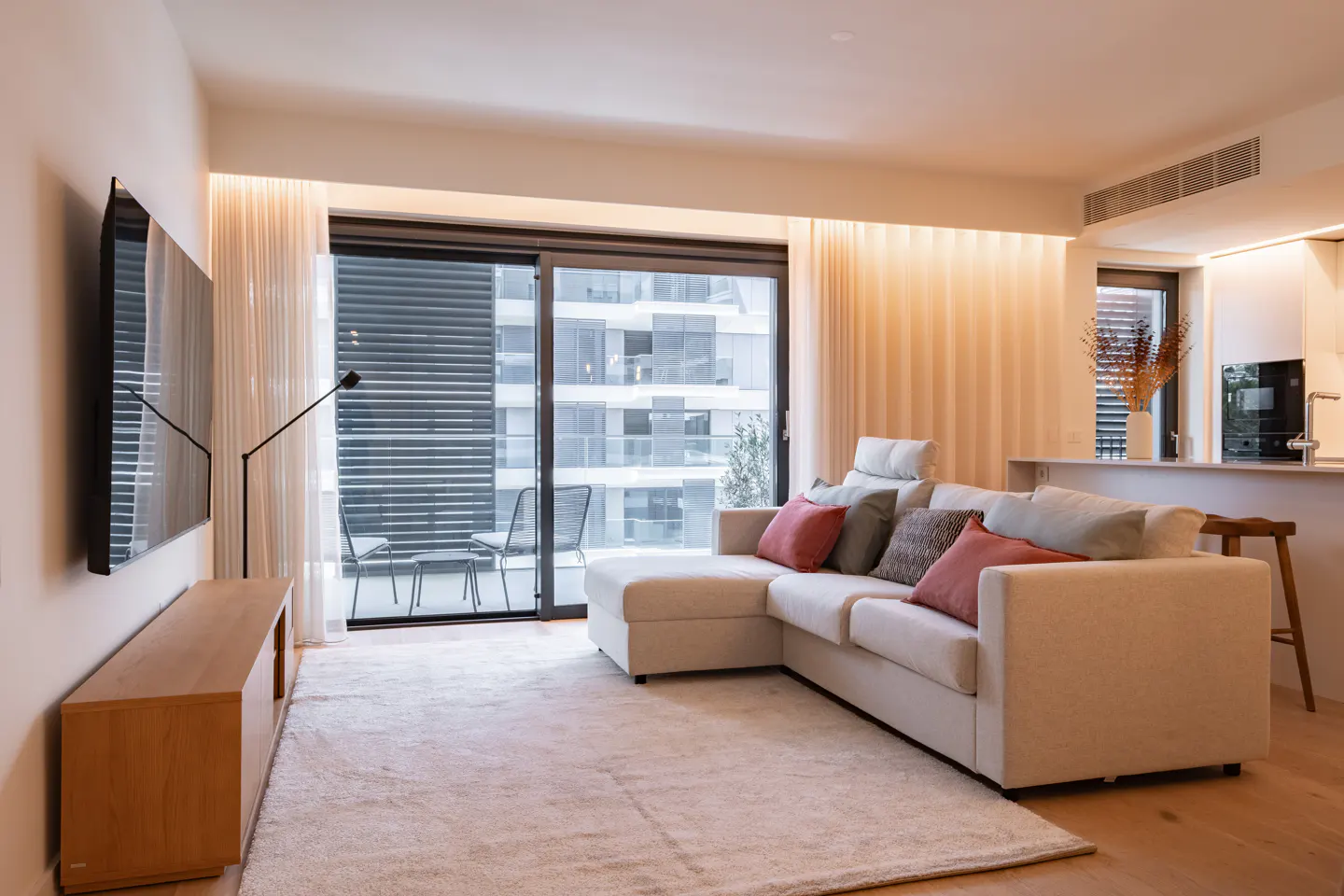 Modern living room with a beige sectional sofa, colorful throw pillows, wooden media console, and balcony view.