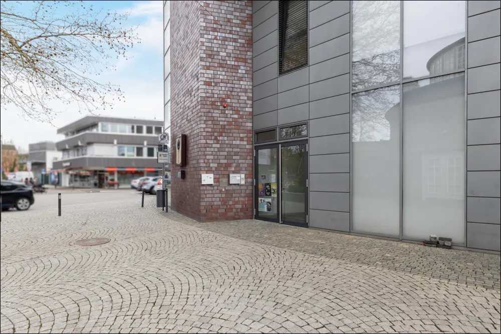 Modern building entrance with brick and grey metal facade, glass door, and cobblestone plaza in a city setting.