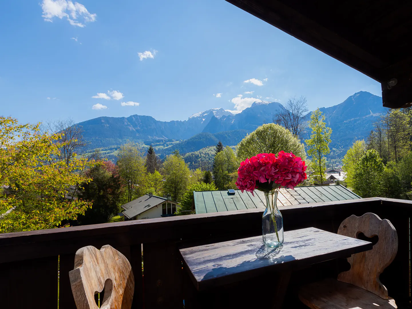 Balcony view with red flowers in a vase on a table, wooden chairs, and mountains in the background on a sunny day.
