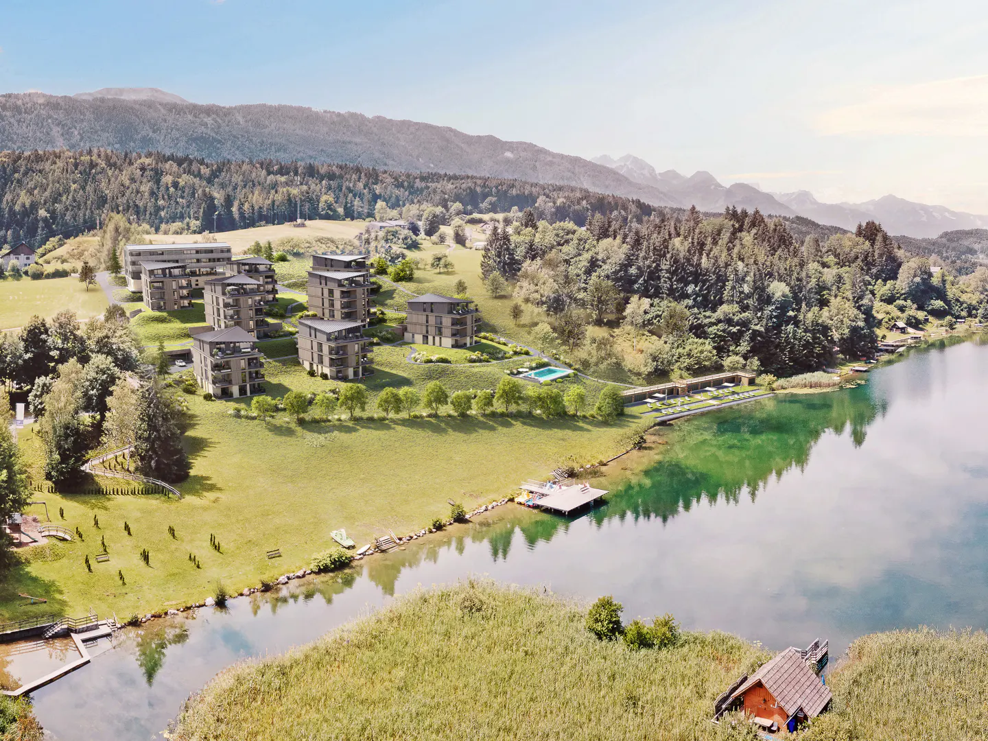 Aerial view of modern apartments on a green hillside near a lake, surrounded by trees and mountains in the background.