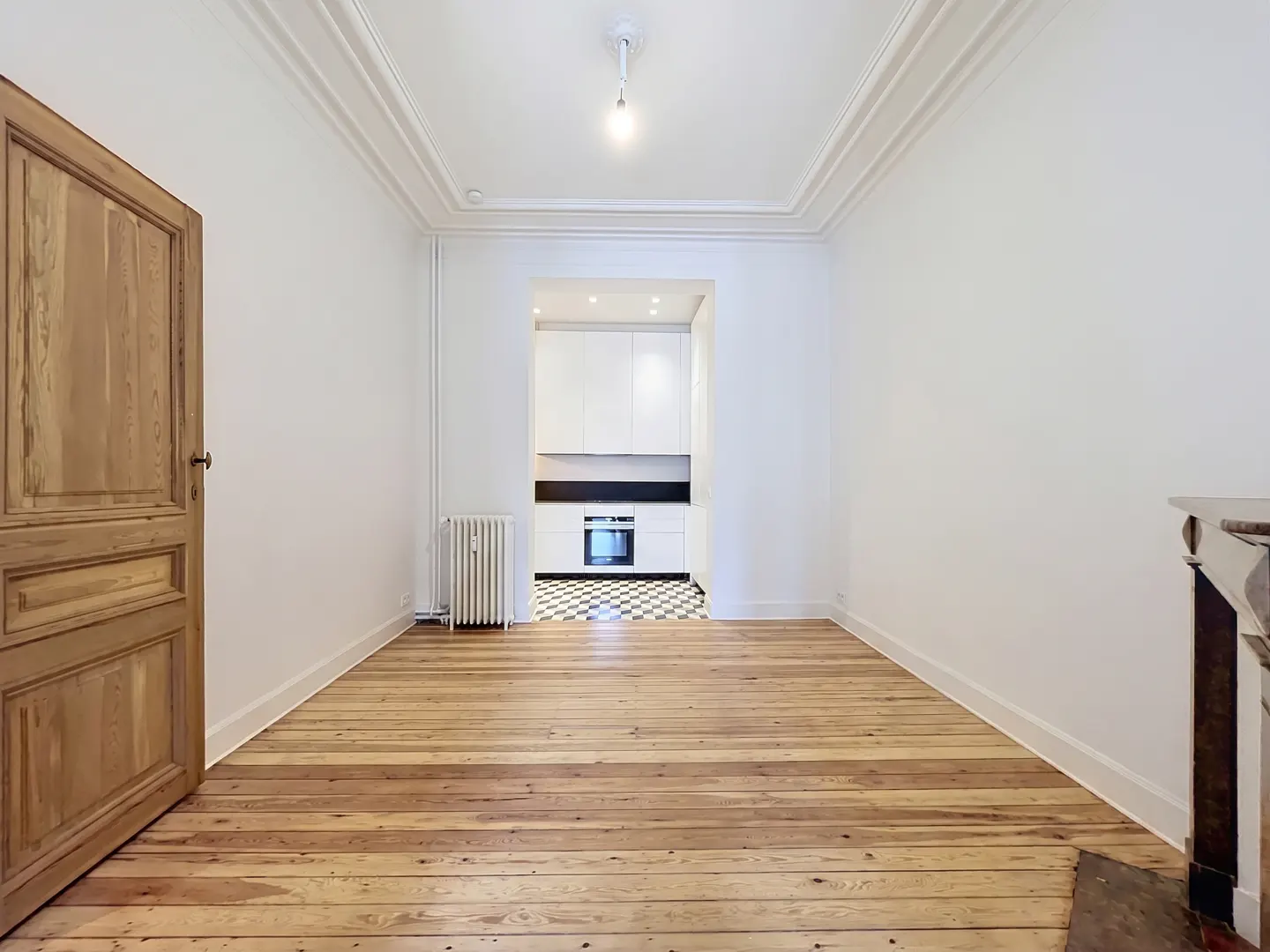 Bright, empty room with wood floors, white walls, and an open doorway to a white kitchen with black and white checkered floors.