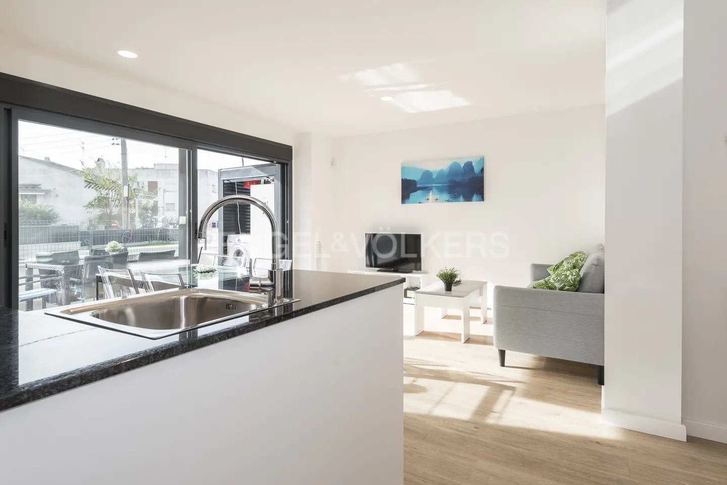 Open-concept living space with a kitchen island, stainless steel sink, gray sofa, and a blue landscape painting on the white wall.